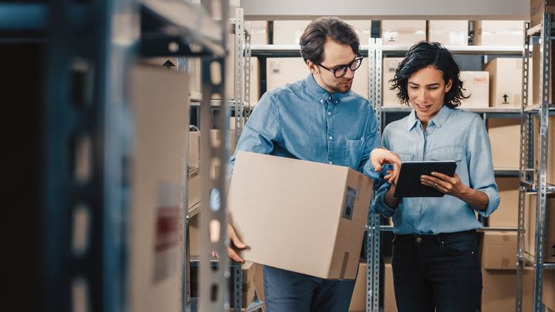 Female Inventory Manager Shows Digital Tablet Information to a Worker Holding Cardboard Box, They Talk and Do Work. In the Background Stock of Parcels with Products Ready for Shipment. Female Inventory Manager Shows Digital Tablet Information to a Worker Holding Cardboard Box, They Talk and Do Work. In the Background Stock of Parcels with Products Ready for Shipment.